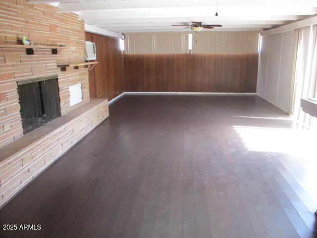 unfurnished living room with dark wood-style floors, beam ceiling, a ceiling fan, wood walls, and a stone fireplace