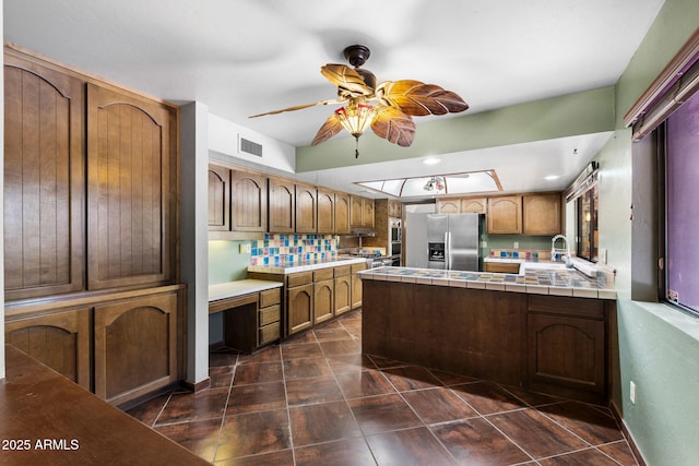 kitchen with stainless steel fridge with ice dispenser, tile countertops, ceiling fan, a peninsula, and a sink