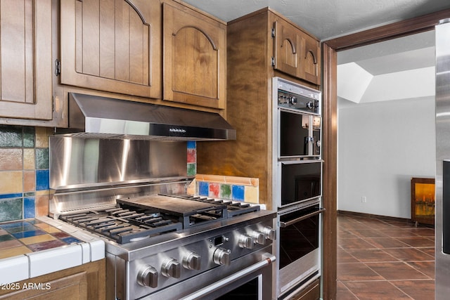 kitchen with tile counters, under cabinet range hood, backsplash, brown cabinetry, and dark tile patterned floors