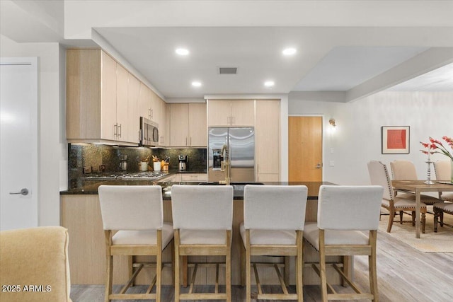 kitchen featuring a breakfast bar area, appliances with stainless steel finishes, light brown cabinetry, decorative backsplash, and light wood-type flooring