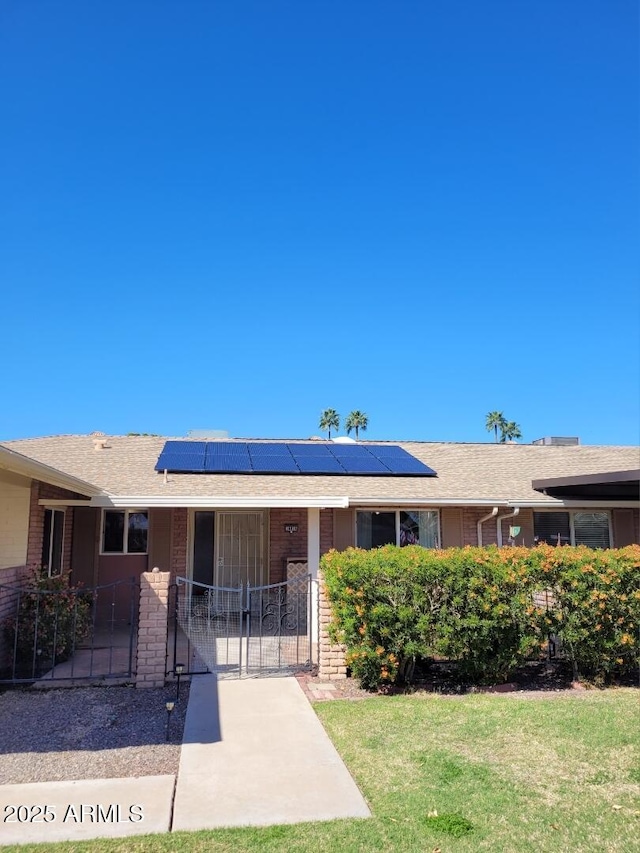 ranch-style house with brick siding, a shingled roof, a front lawn, roof mounted solar panels, and a gate