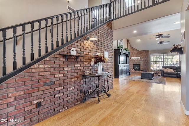 living room with light wood-type flooring, high vaulted ceiling, a brick fireplace, ceiling fan, and brick wall