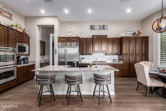 kitchen featuring built in appliances, a center island with sink, dark brown cabinetry, and dark wood-type flooring