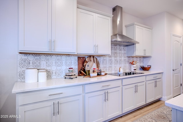 kitchen with wall chimney range hood, white cabinets, black electric cooktop, light wood-type flooring, and backsplash