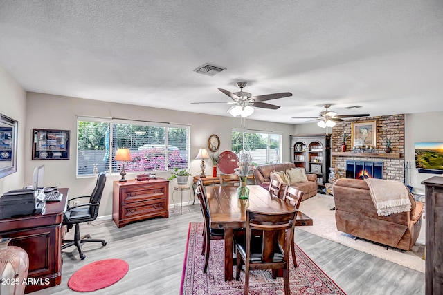 dining room with visible vents, a brick fireplace, ceiling fan, light wood-style floors, and a textured ceiling
