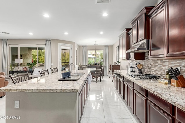 kitchen featuring stainless steel gas cooktop, under cabinet range hood, a center island with sink, decorative backsplash, and marble finish floor