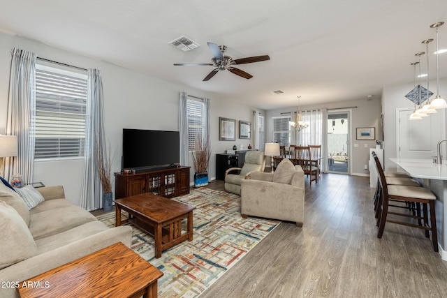 living room featuring light wood-type flooring, visible vents, baseboards, and ceiling fan with notable chandelier