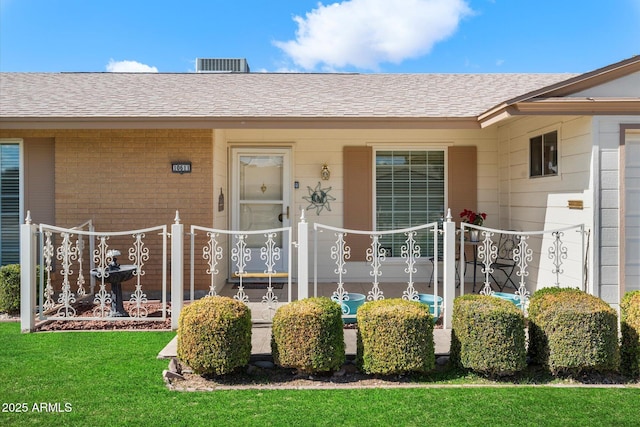 entrance to property featuring a shingled roof, brick siding, a yard, and fence