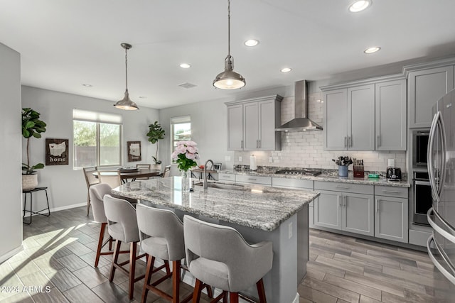 kitchen featuring wall chimney exhaust hood, sink, decorative light fixtures, gray cabinets, and a center island with sink