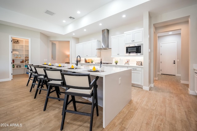 kitchen with stainless steel microwave, wall chimney exhaust hood, an island with sink, a breakfast bar, and white cabinets