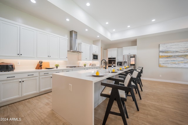 kitchen with appliances with stainless steel finishes, a kitchen island with sink, wall chimney range hood, white cabinets, and light hardwood / wood-style floors