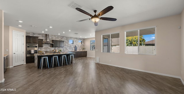 kitchen featuring dark hardwood / wood-style flooring, dark brown cabinetry, a kitchen island with sink, wall chimney range hood, and a breakfast bar area