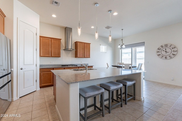 kitchen featuring wall chimney exhaust hood, sink, light tile patterned floors, decorative light fixtures, and a center island with sink