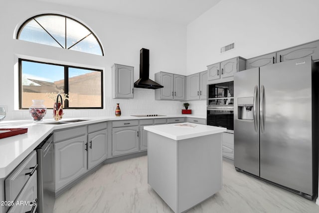 kitchen with gray cabinetry, a high ceiling, black appliances, a kitchen island, and wall chimney exhaust hood