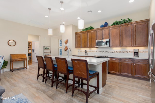 kitchen with an island with sink, stainless steel appliances, decorative light fixtures, tasteful backsplash, and light wood-type flooring