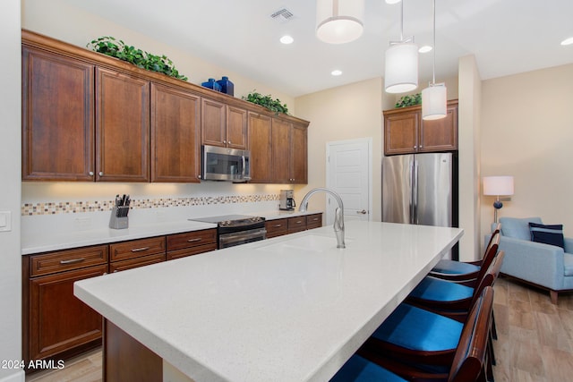 kitchen with appliances with stainless steel finishes, an island with sink, light wood-type flooring, pendant lighting, and backsplash