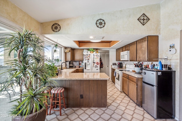 kitchen featuring white appliances, a peninsula, a tray ceiling, a sink, and a breakfast bar area