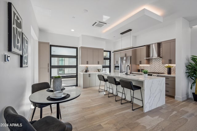 kitchen with a center island with sink, appliances with stainless steel finishes, light wood-type flooring, wall chimney range hood, and pendant lighting