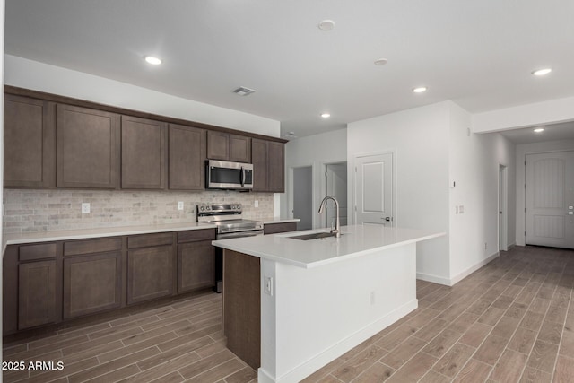 kitchen with sink, appliances with stainless steel finishes, dark brown cabinets, a center island with sink, and decorative backsplash