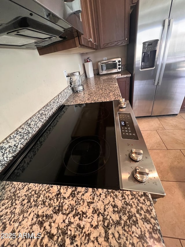 kitchen featuring stainless steel fridge, cooktop, light tile patterned flooring, dark stone countertops, and dark brown cabinets