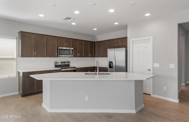 kitchen featuring sink, appliances with stainless steel finishes, a kitchen island with sink, dark brown cabinets, and tasteful backsplash