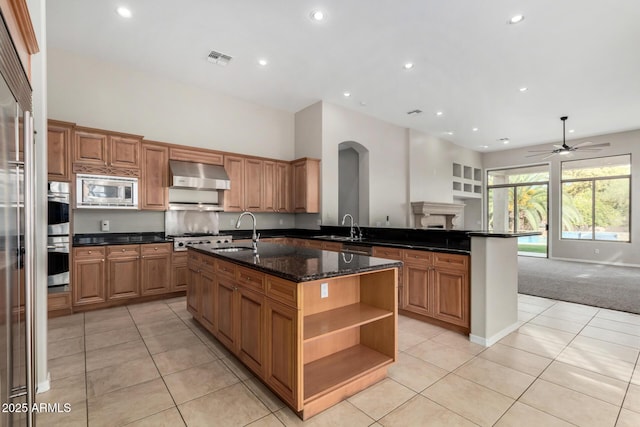 kitchen with wall chimney range hood, sink, appliances with stainless steel finishes, dark stone countertops, and an island with sink