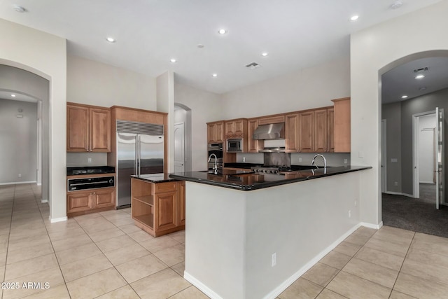 kitchen featuring wall chimney exhaust hood, built in appliances, a center island with sink, light tile patterned floors, and kitchen peninsula