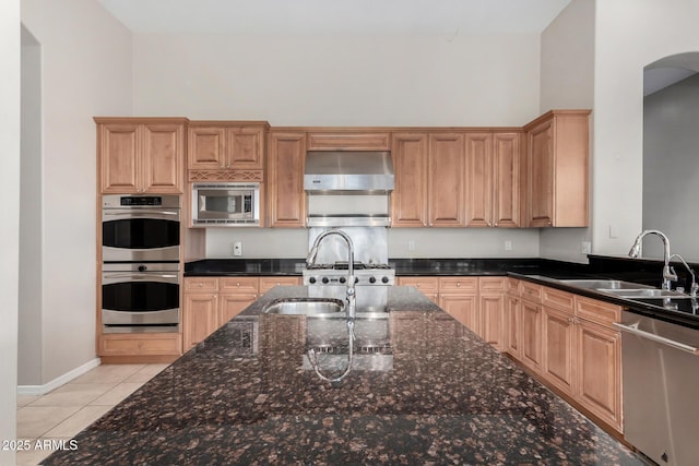 kitchen with light tile patterned floors, sink, range hood, stainless steel appliances, and dark stone counters