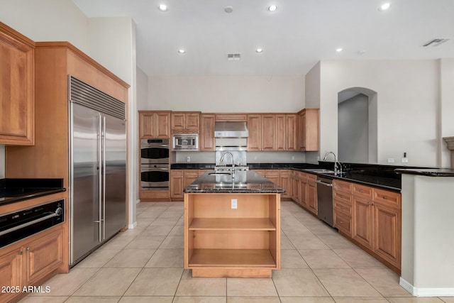 kitchen with light tile patterned floors, sink, built in appliances, extractor fan, and an island with sink