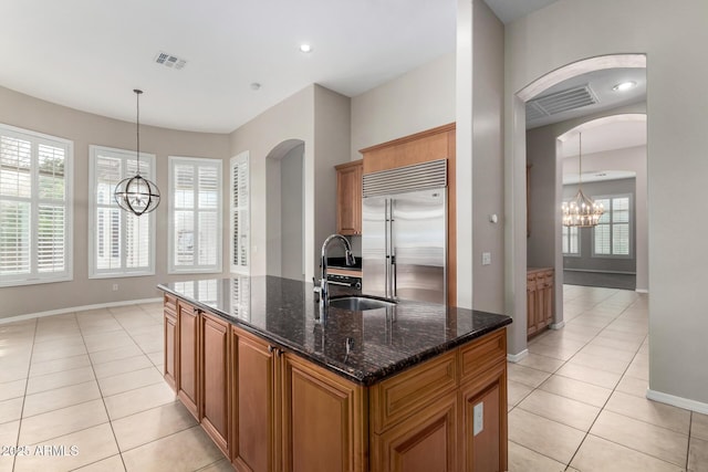 kitchen featuring built in fridge, pendant lighting, an island with sink, sink, and dark stone counters