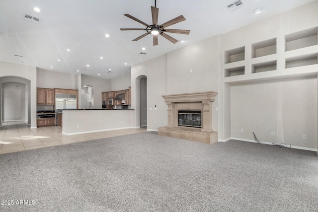 unfurnished living room featuring a tiled fireplace, built in shelves, light colored carpet, and ceiling fan