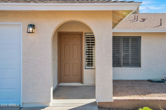 view of exterior entry with a garage, stucco siding, and roof with shingles