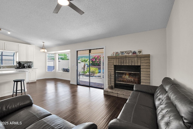 living area with dark wood-type flooring, vaulted ceiling, a fireplace, a textured ceiling, and a ceiling fan