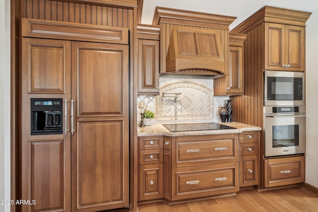 kitchen with light wood-style flooring, light stone counters, brown cabinets, custom exhaust hood, and stainless steel appliances