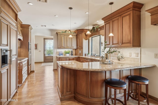 kitchen featuring tasteful backsplash, brown cabinetry, light stone countertops, light wood-type flooring, and a peninsula