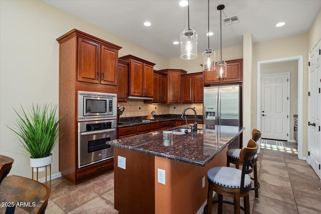 kitchen with an island with sink, sink, dark stone countertops, hanging light fixtures, and stainless steel appliances