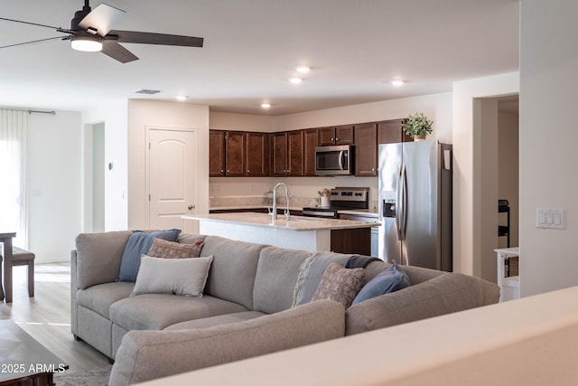 kitchen featuring dark brown cabinets, a center island with sink, ceiling fan, stainless steel appliances, and light hardwood / wood-style floors