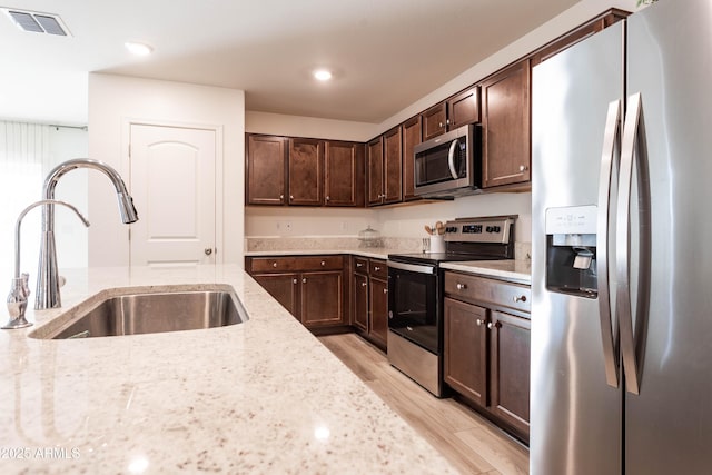 kitchen with sink, appliances with stainless steel finishes, light stone counters, dark brown cabinetry, and light hardwood / wood-style floors