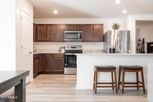 kitchen with light stone countertops, appliances with stainless steel finishes, dark brown cabinetry, and light hardwood / wood-style flooring