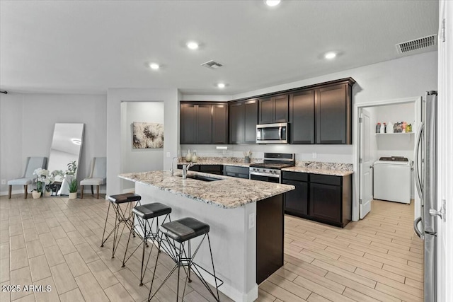 kitchen with sink, light stone countertops, washer / dryer, a kitchen island with sink, and appliances with stainless steel finishes