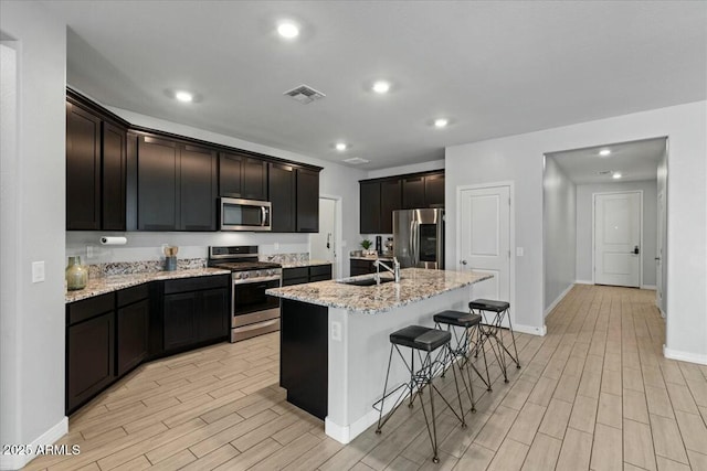 kitchen with light stone counters, stainless steel appliances, a kitchen island with sink, a breakfast bar area, and sink