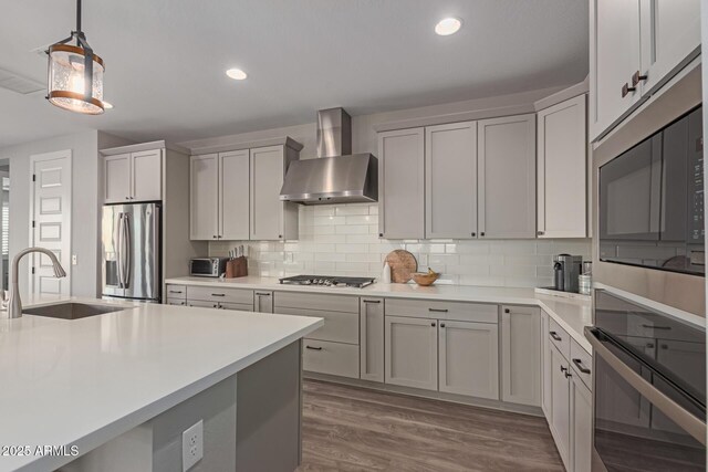 kitchen featuring stainless steel appliances, light countertops, hanging light fixtures, a sink, and wall chimney range hood