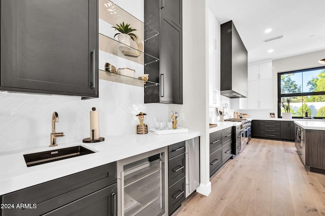 kitchen featuring light wood-style flooring, wine cooler, a sink, wall chimney range hood, and backsplash