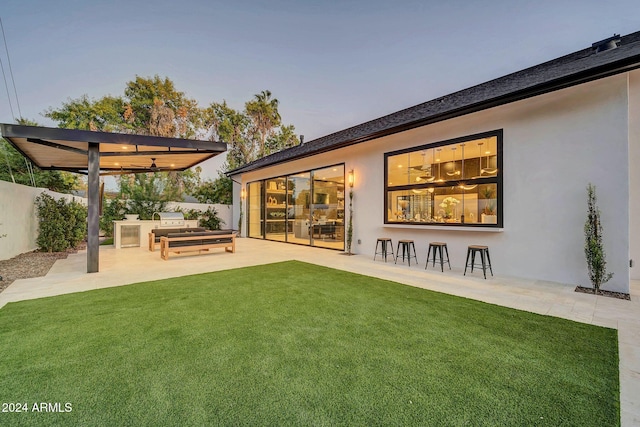 back house at dusk featuring a patio, ceiling fan, a lawn, and exterior kitchen