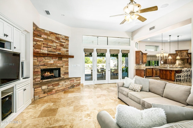 living room with visible vents, a high ceiling, a stone fireplace, and ceiling fan with notable chandelier