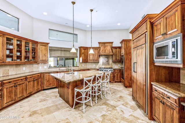 kitchen with light stone counters, a breakfast bar, an island with sink, built in appliances, and brown cabinets