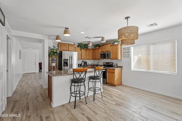 kitchen with light hardwood / wood-style flooring, appliances with stainless steel finishes, a kitchen island with sink, hanging light fixtures, and a kitchen bar