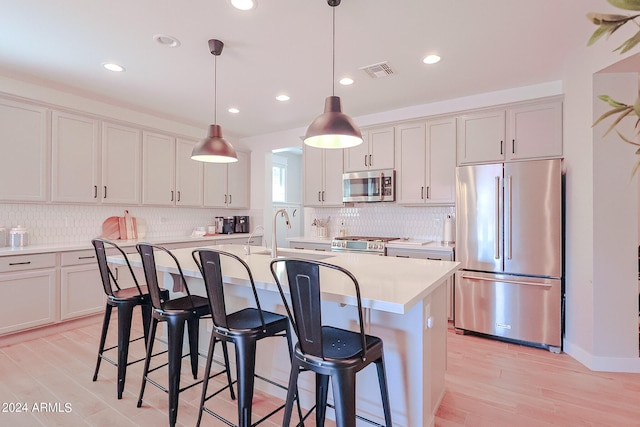 kitchen featuring appliances with stainless steel finishes, hanging light fixtures, decorative backsplash, light wood-type flooring, and a kitchen island with sink