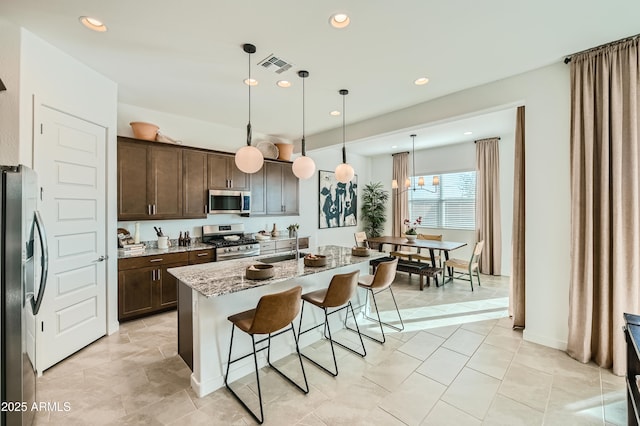 kitchen with visible vents, light stone counters, stainless steel appliances, dark brown cabinets, and a kitchen bar