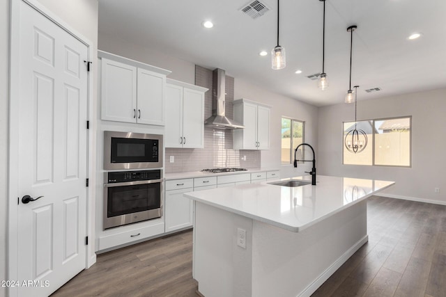 kitchen with wall chimney exhaust hood, stainless steel appliances, sink, decorative light fixtures, and an island with sink